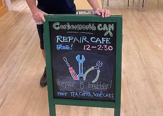 Person standing beside a sign for Cockermouth CAN's repair cafe